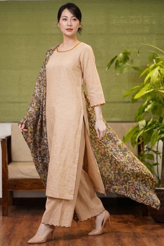 Full-length view of a model walking in the beige Sand Dot Kurti set, featuring a straight-cut silhouette with small dot motifs, matching wide-leg trousers with scalloped hems, and a dark, flowing floral printed dupatta.