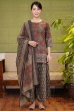 Full-length, front-facing view of a model wearing the complete Sepia Halo Kurti set. The image captures the rich contrast of red circular block prints against the warm sepia base of the kurti, coordinated flawlessly with the distinct botanical pattern on the flared bottom and the wide borders of the draped dupatta. The look is set against a premium minimal studio background featuring a wooden sofa with beige cushions and an indoor plant.