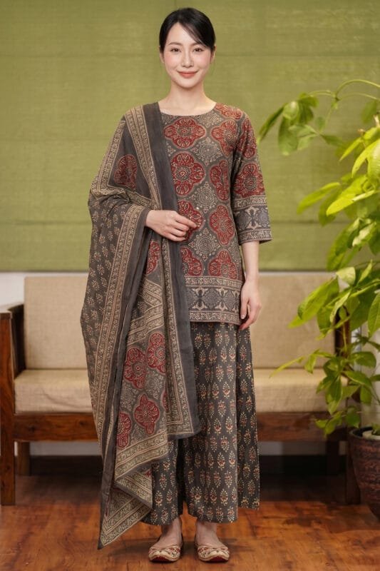 Full-length, front-facing view of a model wearing the complete Sepia Halo Kurti set. The image captures the rich contrast of red circular block prints against the warm sepia base of the kurti, coordinated flawlessly with the distinct botanical pattern on the flared bottom and the wide borders of the draped dupatta. The look is set against a premium minimal studio background featuring a wooden sofa with beige cushions and an indoor plant.