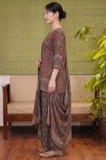 Full-length side profile view of a model wearing the Sepia Halo Kurti set, emphasizing the straight-cut hem and intricate red circular motifs. The look is paired with matching flared bottoms showcasing repeating botanical prints and a beautifully draped patterned dupatta, captured in a standardized studio setting with warm wooden accents and greenery.