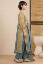 Full-length side profile of a model wearing the Ziya Notch Kurti set, showcasing the straight-cut hem and matching Dusty Blue straight trousers. The draped Dusty Blue dupatta features a delicate golden scalloped lace border, set against a minimal studio background with a cane-weaved wooden divider.