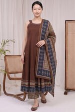 A full-length view of a woman wearing a brown sleeveless Ajrakh Kurti featuring vertical pleats, paired with a patterned dupatta and matching printed trousers in a minimal studio setting.