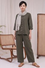A woman wearing the Sage Coord in olive green, featuring a button-down shirt with a collar, matching wide-leg trousers, and a patterned scarf in a minimal studio.