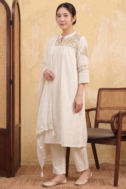A woman posing in a full-length ivory Zari Kurti with intricate gold embroidery, matching straight trousers, and a textured dupatta, set against a minimal studio background with cane-weaved wooden furniture.