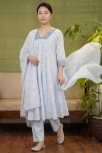 Full-length shot of a model wearing the complete Blossom Blue Chanderi Kurti Set in a dusty blue color. The ensemble features a gathered kurti, perfectly matched pants, and a beautifully draped dupatta, emphasizing the standardized yoke pattern and cohesive hem detailing. Set against a premium background with minimal wooden elements and a cane-weaved chair.