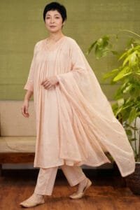 Full-length shot of a model wearing the complete Chanderi Blossom Cream Elegance Kurti set. The ensemble features the gathered cream kurti, matched straight pants, and an elegantly draped dupatta, emphasizing the standardized yoke pattern and cohesive styling. Set against a premium background with minimal wooden furniture and an indoor plant.