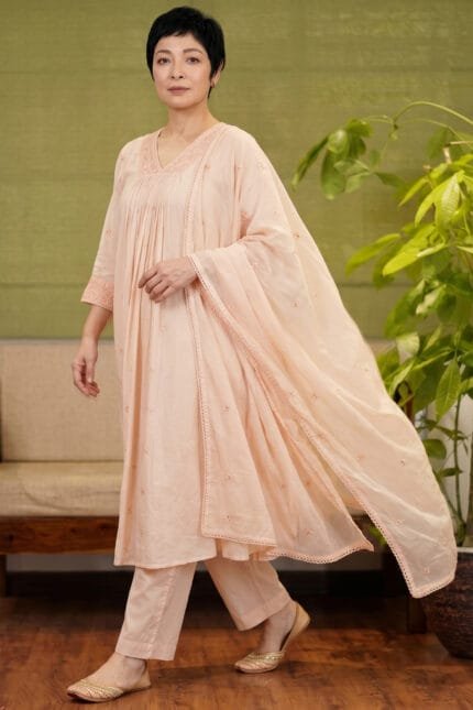 Full-length shot of a model wearing the complete Chanderi Blossom Cream Elegance Kurti set. The ensemble features the gathered cream kurti, matched straight pants, and an elegantly draped dupatta, emphasizing the standardized yoke pattern and cohesive styling. Set against a premium background with minimal wooden furniture and an indoor plant.