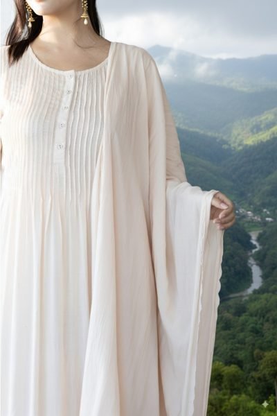A woman wearing a soft white pleated kurti with dupatta, standing against the scenic hills and valleys of Mizoram, highlighting elegant ethnic fashion.